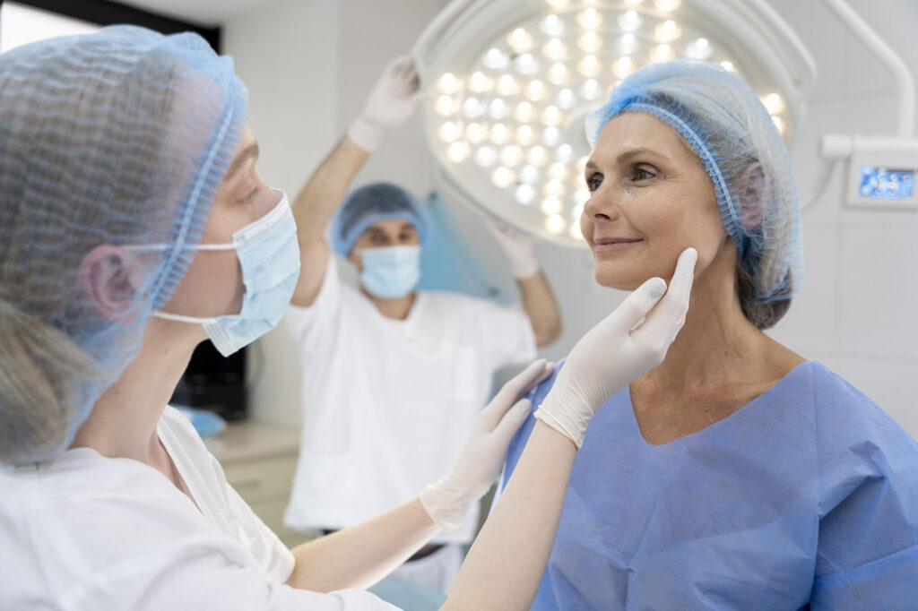 A medical professional examines a woman's face in a clinic, both wearing surgical caps and masks, whilst another staff member adjusts an overhead light.