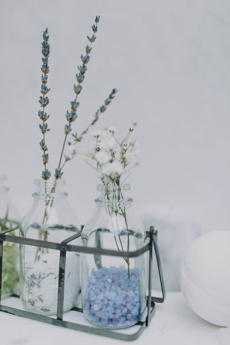 Three glass bottles in a metal holder—one with lavender, one with white flowers, and one with blue bath salts—bring a calming touch, ideal for an OCD Clinic Brisbane setting, set against a light background.