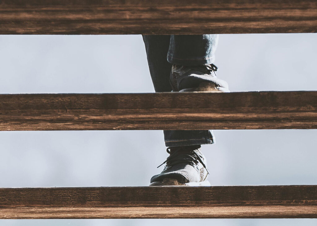 Close-up of a person’s legs and feet wearing dark shoes and jeans ascending wooden stairs, viewed from below.
