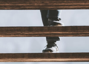 Close-up of a person’s legs and feet wearing dark shoes and jeans ascending wooden stairs, viewed from below.